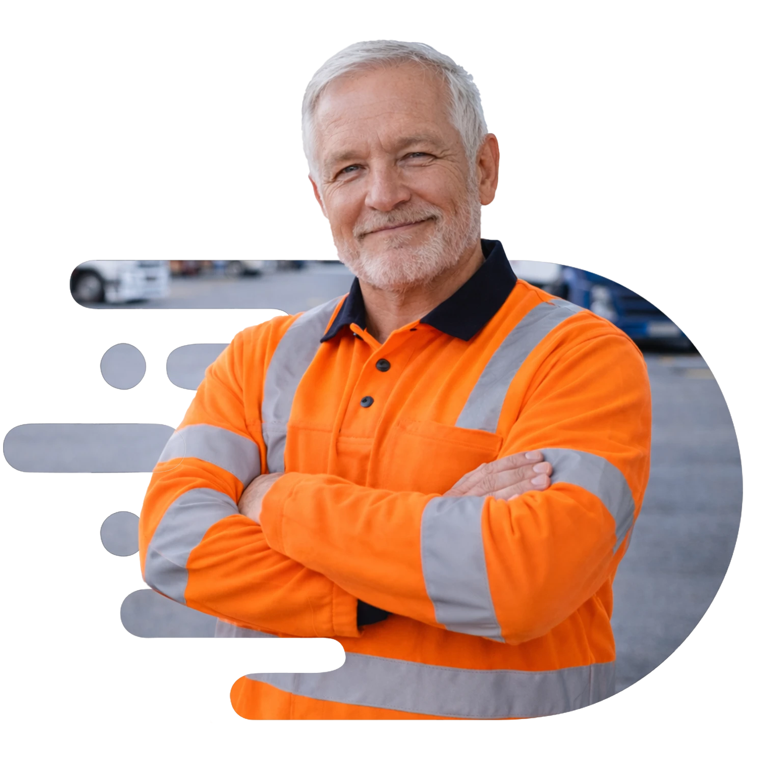 Older Caucasian truck driver in a long-sleeve orange high-visibility shirt standing with arms crossed in a trucking yard, smiling at the camera with parked trucks softly blurred in the background.